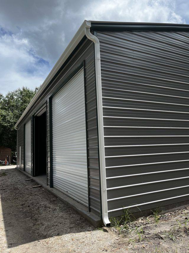 White gutter and downspout on the corner of a dark gray metal building with roll-up doors