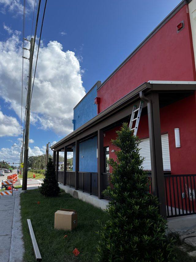A ladder against the porch roof of a red and blue storefront, with a downspout section waiting on the grass