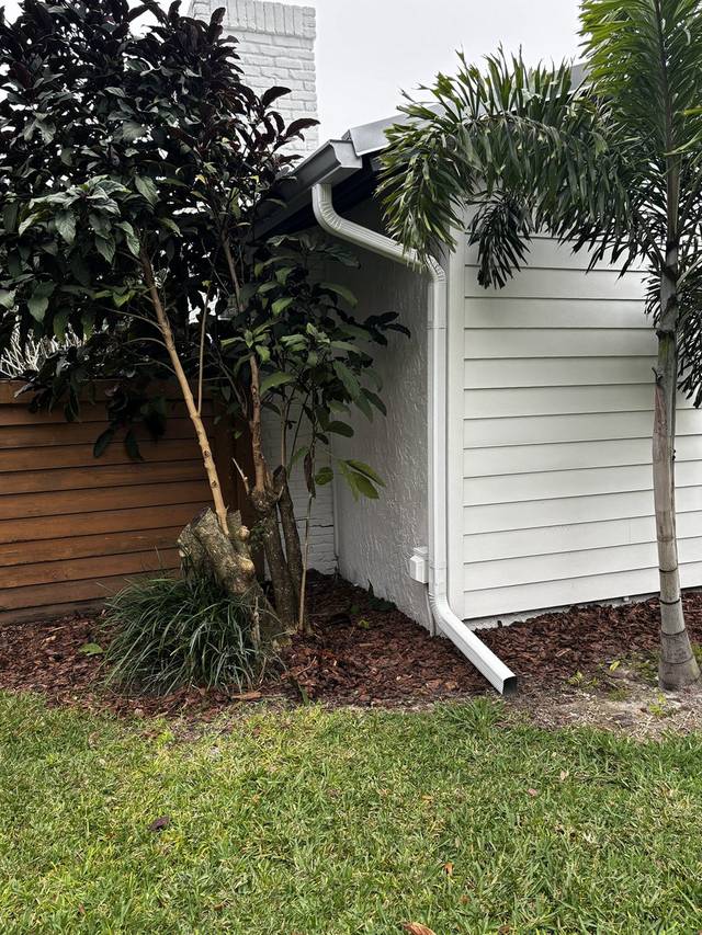 White downspout with a short extension on the side of a white home, beside a palm and a wood fence